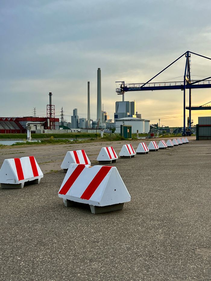 A row of red and white barriers at an industrial port with cranes and factories at sunset.